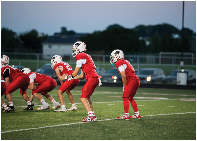 Several people in red football uniforms and white helmets are positioned on a football field, preparing for a play during a game at dusk. Description generated by AI