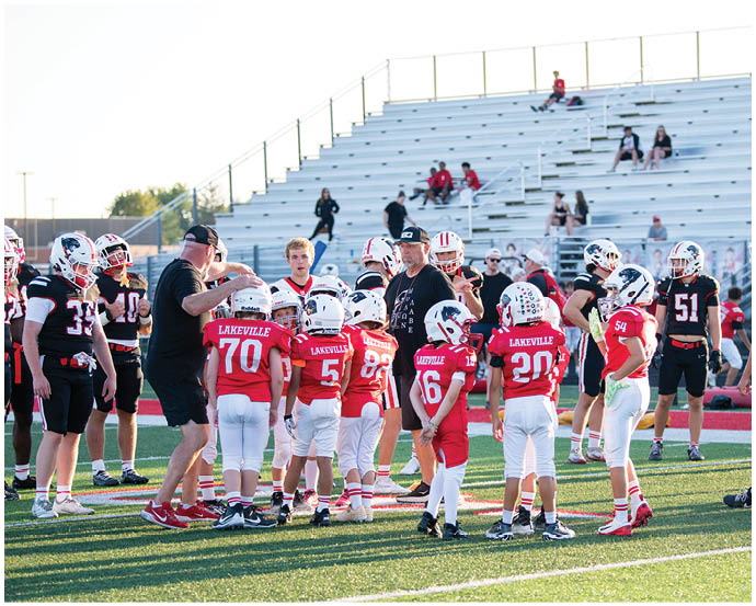 A group of children in red and white football uniforms are gathered on a field with adults in black coaching them while other players and people sit in the bleachers in the background. Description generated by AI