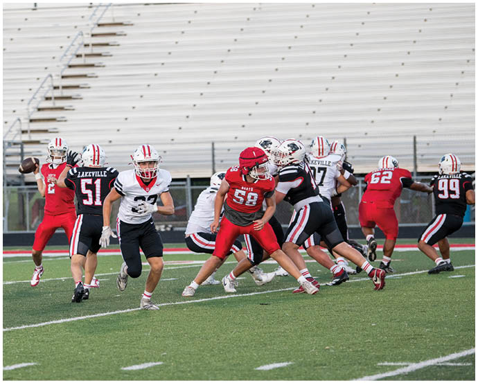 Players in red and black football uniforms are engaged in a play on a green field with empty bleachers in the background. Description generated by AI