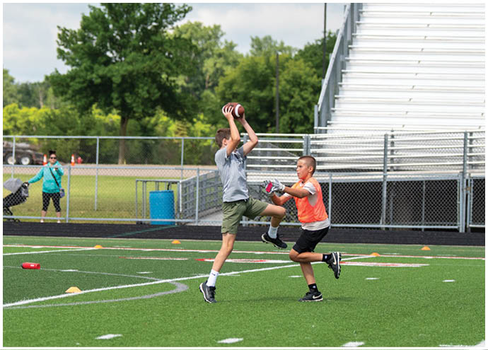 Two people wearing athletic clothing are playing a sport on a green field with one person jumping and the other reaching out with gloved hands. Description generated by AI