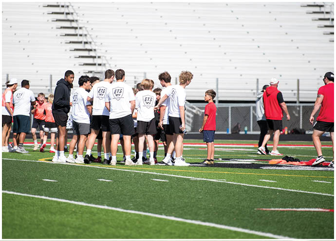 A group of people wearing white shirts and shorts are gathered on a sports field near a few others in red shirts, with empty bleachers in the background. Description generated by AI