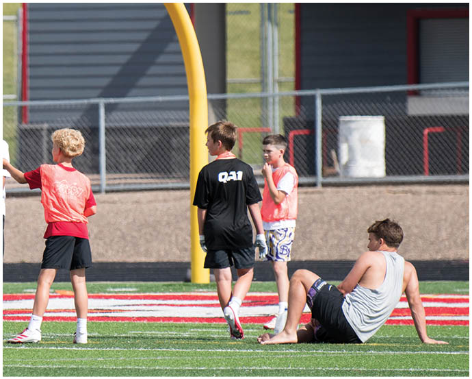 A group of people wearing casual sportswear are gathered on a football field near a goalpost, with one person sitting on the ground and others standing and interacting. Description generated by AI