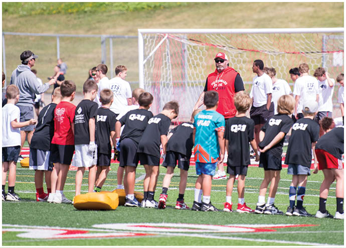 A group of children in sportswear stand in a line on a soccer field facing adults near a goalpost, with one adult in a red hoodie appearing to give instructions. Description generated by AI