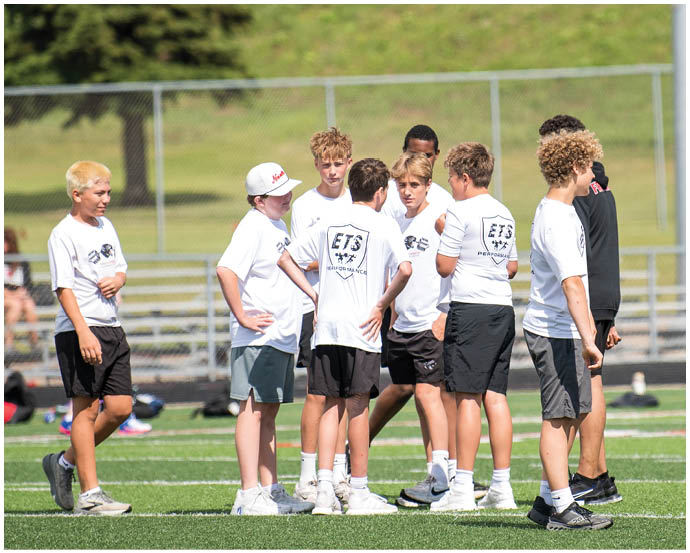 A group of children wearing white sports shirts and shorts are standing and talking on a grassy sports field with a chain-link fence and bleachers in the background. Description generated by AI