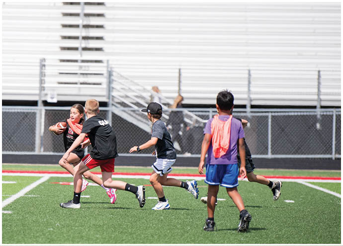Children playing a game of flag football on a green field with bleachers in the background. Description generated by AI