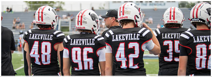 Five people wearing black sports uniforms with red and white stripes stand on a field with their backs facing the camera. Description generated by AI