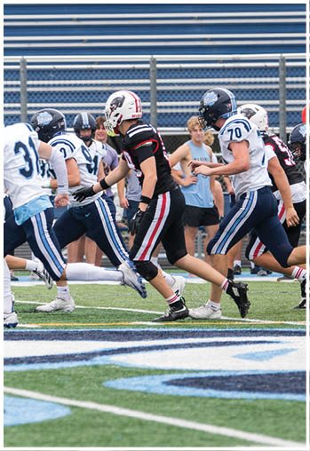People in football uniforms are running on a field during a game with spectators in the background. Description generated by AI