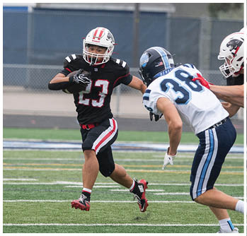 A person wearing a black football uniform with the number 23 is running with the ball while another person in a white and blue uniform with the number 38 is approaching on a football field. Description generated by AI