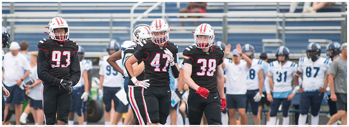 Four football players in black uniforms with red and white helmets stand on a field with a crowd and other players in white uniforms in the background. Description generated by AI