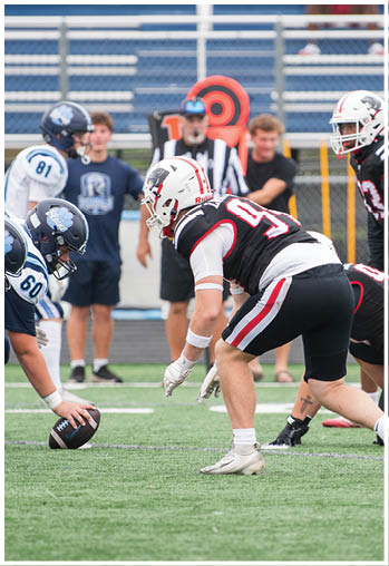 Two people in football uniforms are crouched on a field preparing for a play while others and a referee stand in the background near bleachers. Description generated by AI