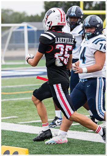 A person wearing a black football uniform and helmet is running on a football field near a yellow marker indicating the 20-yard line. Description generated by AI