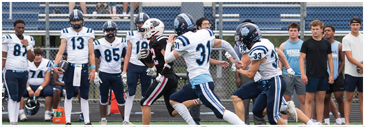 People in football uniforms are actively playing on a field while others in casual clothing watch from the sidelines and bleachers. Description generated by AI