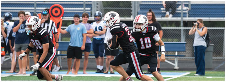 Three people in black football uniforms are actively playing on a field while several people watch from the sidelines. Description generated by AI