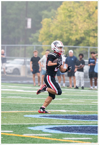 A person in a black football uniform with red and white stripes is running on a field with a helmet on while several people stand in the background near a fence. Description generated by AI