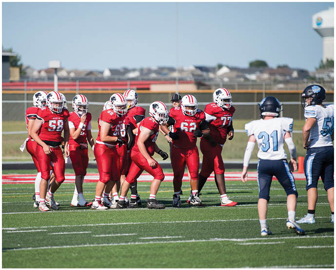 A group of people in red football uniforms are gathered on a field facing two people in white and blue football uniforms. Description generated by AI