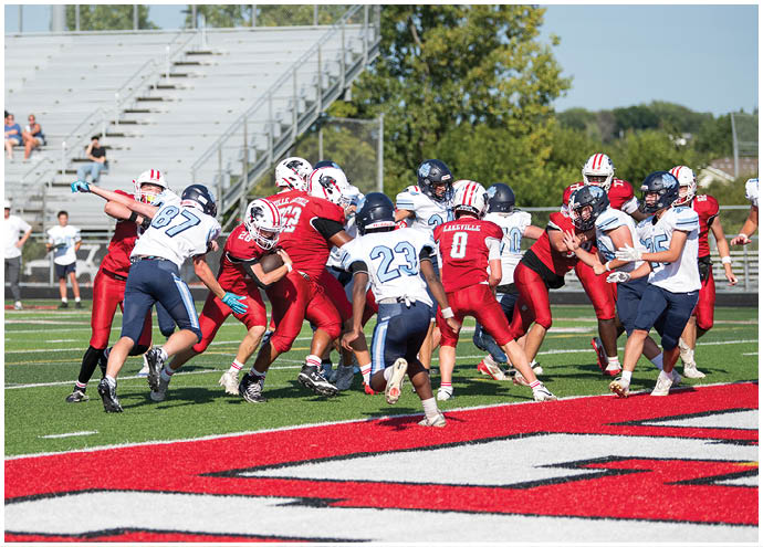 A group of people wearing red and white and blue and white football uniforms are actively playing on a green field near the end zone with red and white markings. Description generated by AI