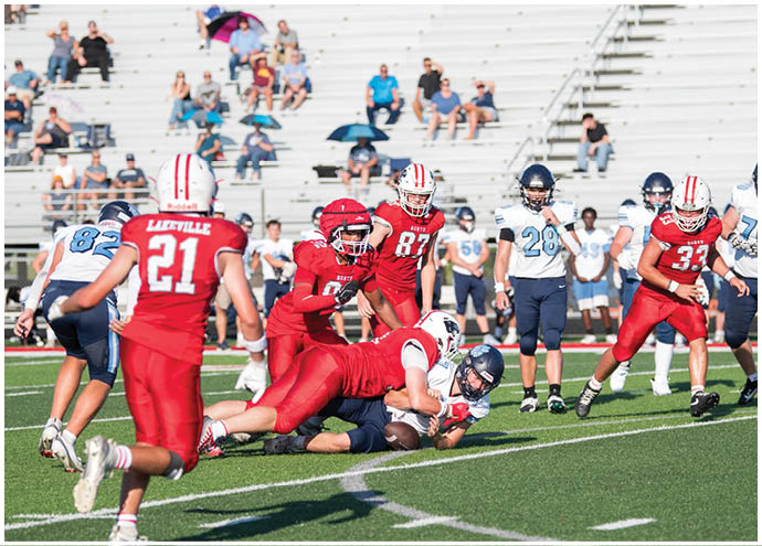 Players in red and white uniforms tackle a player in a white and blue uniform during a football game on a green field with spectators in the background. Description generated by AI