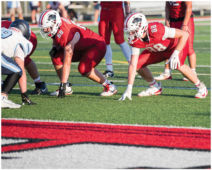 Two football players in red uniforms are crouched on a green field facing two players in white and blue uniforms, preparing for a play. Description generated by AI