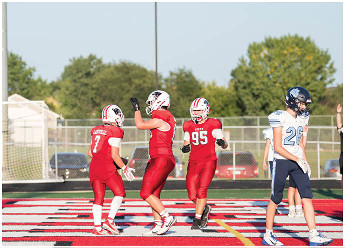 Four people in football uniforms stand on a field with a red and white patterned end zone, with three wearing red and one wearing white and blue. Description generated by AI