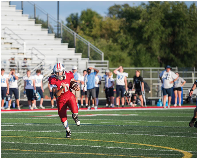 A person in a red football uniform is running with the ball on a field while a referee runs nearby and people stand in the background. Description generated by AI