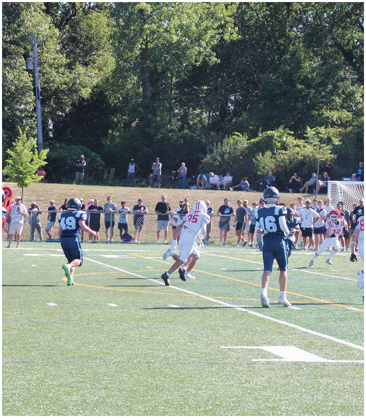Adults in sports uniforms are playing a football game on a grassy field with spectators watching from the sidelines and a wooded area in the background. Description generated by AI