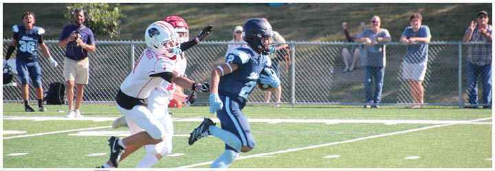 Two people in football uniforms are running on a field with a chain-link fence and spectators in the background. Description generated by AI