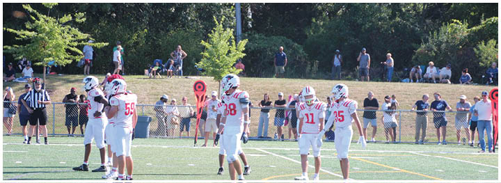 People in football uniforms stand on a field while referees and spectators watch from the sidelines and a grassy hill in the background. Description generated by AI
