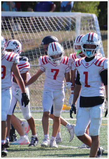 Several people wearing white and red football uniforms are gathered on a field near a goalpost, with one person in the foreground wearing a helmet and faceguard. Description generated by AI