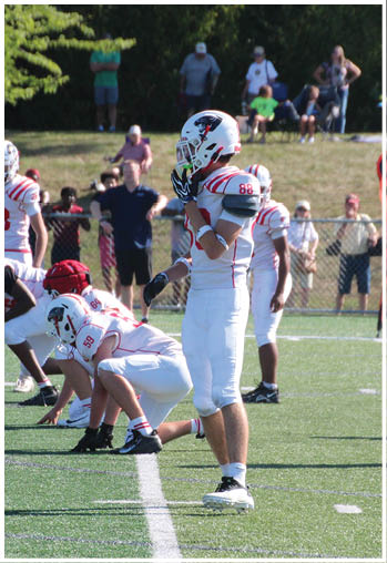 A group of people in football uniforms are positioned on a field with one standing and others crouched, while spectators watch from behind a fence. Description generated by AI