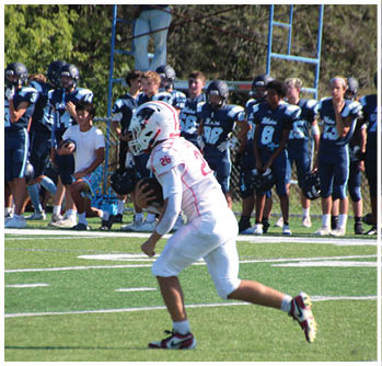 A person in white football gear is running with the ball on a field while a group of people in dark blue uniforms watch from the sidelines. Description generated by AI