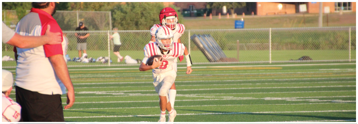 A person in a white and red sports uniform is running on a green football field while another person in casual clothing stands nearby. Description generated by AI