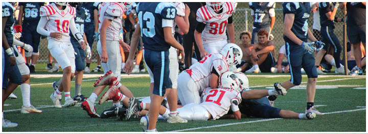 People in football uniforms are on a field with some players standing and others on the ground during a game. Description generated by AI