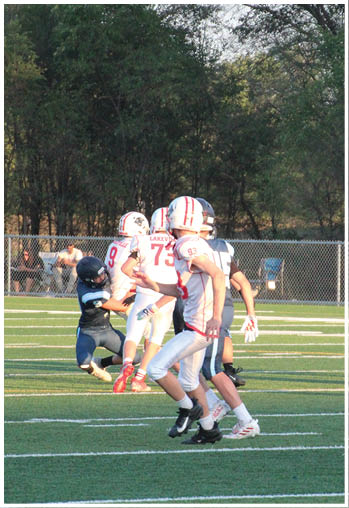 Several people wearing football uniforms are actively playing on a green field with white yard lines and a chain-link fence and trees in the background. Description generated by AI