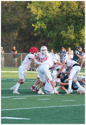 A group of people in white football uniforms are engaged in a play on a green field while others in dark uniforms are on the ground. Description generated by AI