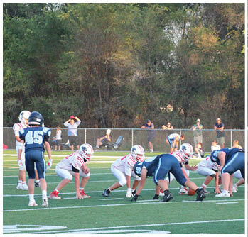 Two teams of youth football players are lined up facing each other on a green field with spectators watching behind a chain-link fence and trees in the background. Description generated by AI