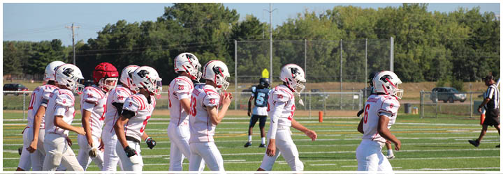 A group of people wearing white and red football uniforms and helmets are walking on a football field with trees and a fence in the background. Description generated by AI