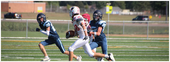 Three people in football uniforms are actively playing on a field with one person holding the ball while the others appear to be defending. Description generated by AI