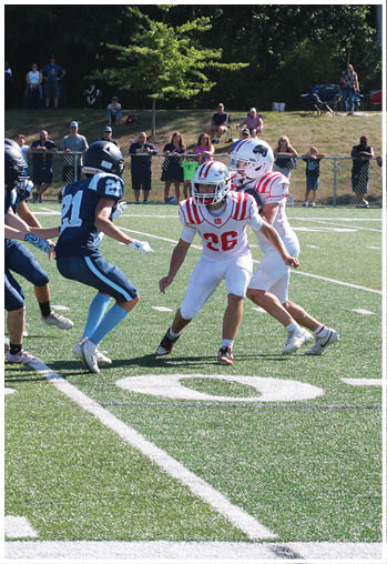 Two people in white and red football uniforms are facing two people in dark blue football uniforms on a green field with spectators in the background. Description generated by AI
