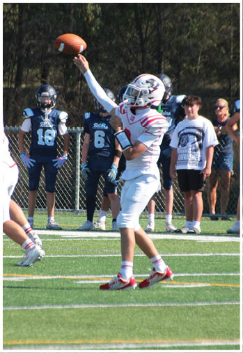 A person in a white and red football uniform is throwing a football while other players in white and dark uniforms stand on a green field with a chain-link fence and trees in the background. Description generated by AI