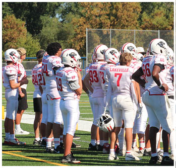 A group of people wearing football uniforms and helmets stand on a field with trees and a fence in the background. Description generated by AI