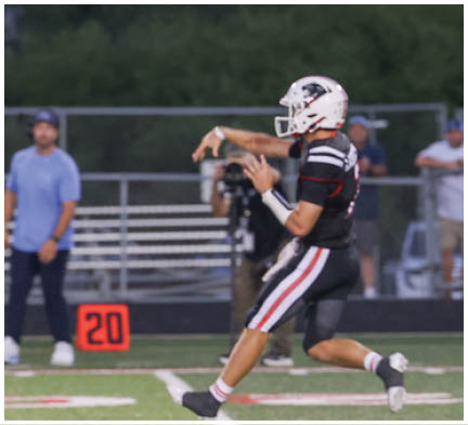 A person wearing a football uniform and helmet is in the motion of throwing a football on a field with a blurred background of people and bleachers. Description generated by AI