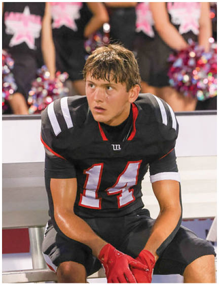 A person wearing a black football uniform with the number 14 and red gloves is sitting on a bench looking to the side with cheerleaders holding pom-poms blurred in the background. Description generated by AI