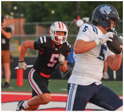 An adult in a white and blue football uniform is holding a football and running on a field while another adult in a black and red uniform chases behind. Description generated by AI
