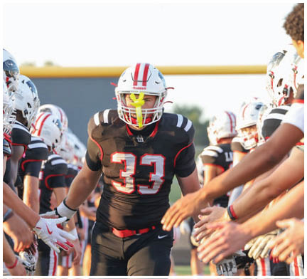 A person wearing a black football uniform with the number 33 is walking between two lines of teammates in white helmets and black uniforms who are reaching out to give high fives. Description generated by AI