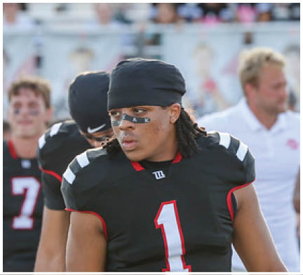A person wearing a black football uniform with the number one and a black head covering stands in front of two other people in similar uniforms on a sports field. Description generated by AI