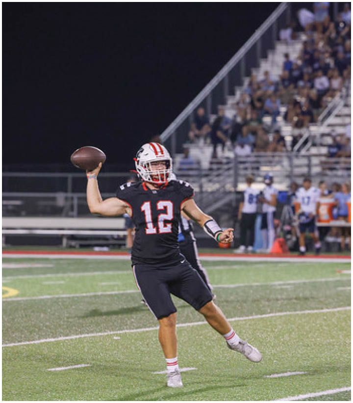 A person wearing a black football uniform with the number 12 is preparing to throw a football on a field at night with spectators in the background. Description generated by AI