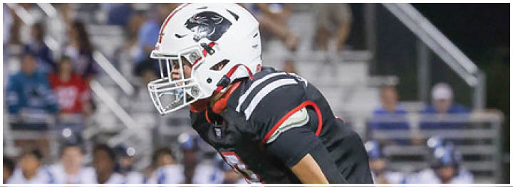A person wearing a black football uniform and white helmet is in a ready stance on a football field with blurred spectators in the background. Description generated by AI