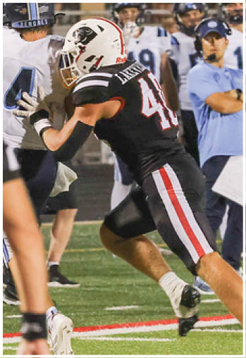 An adult in a black football uniform with the number 48 is tackling or blocking another adult in a white and blue football uniform during a game on a grassy field. Description generated by AI