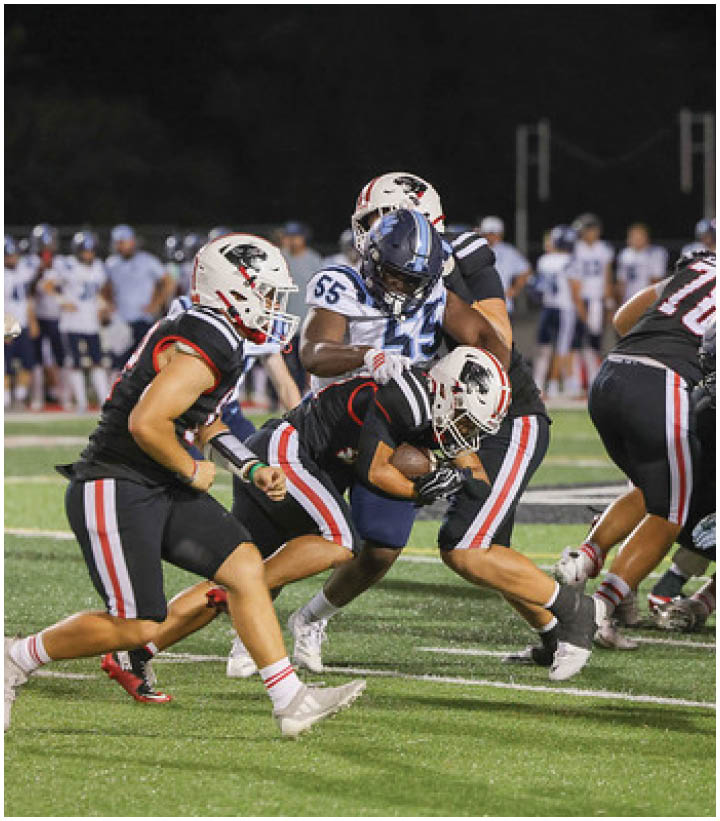 Three football players in black uniforms tackle a player in a white and blue uniform on a field during a nighttime game. Description generated by AI