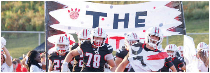A group of people wearing football helmets and uniforms are running through a torn banner that reads “THE" with a paw print symbol above the text. Description generated by AI